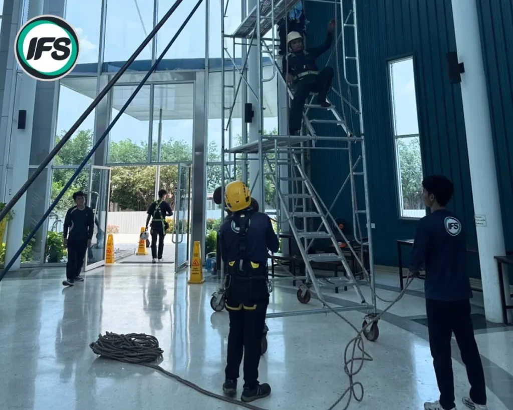 เจ้าหน้าที่ IFS กำลังปฏิบัติการฝึกอบรมความปลอดภัยในการทำงานบนที่สูง ภายในอาคาร โดยมีการสวมใส่อุปกรณ์ป้องกันส่วนบุคคลครบถ้วน ทั้งหมวกนิรภัย สายรัดนิรภัย และอุปกรณ์โรยตัว พร้อมทีมงานประจำจุดคอยดูแลความปลอดภัยอย่างใกล้ชิด บริเวณรอบพื้นที่มีการจัดเก็บอุปกรณ์และติดตั้งสัญลักษณ์แจ้งเตือนเพื่อความปลอดภัยตลอดการฝึก