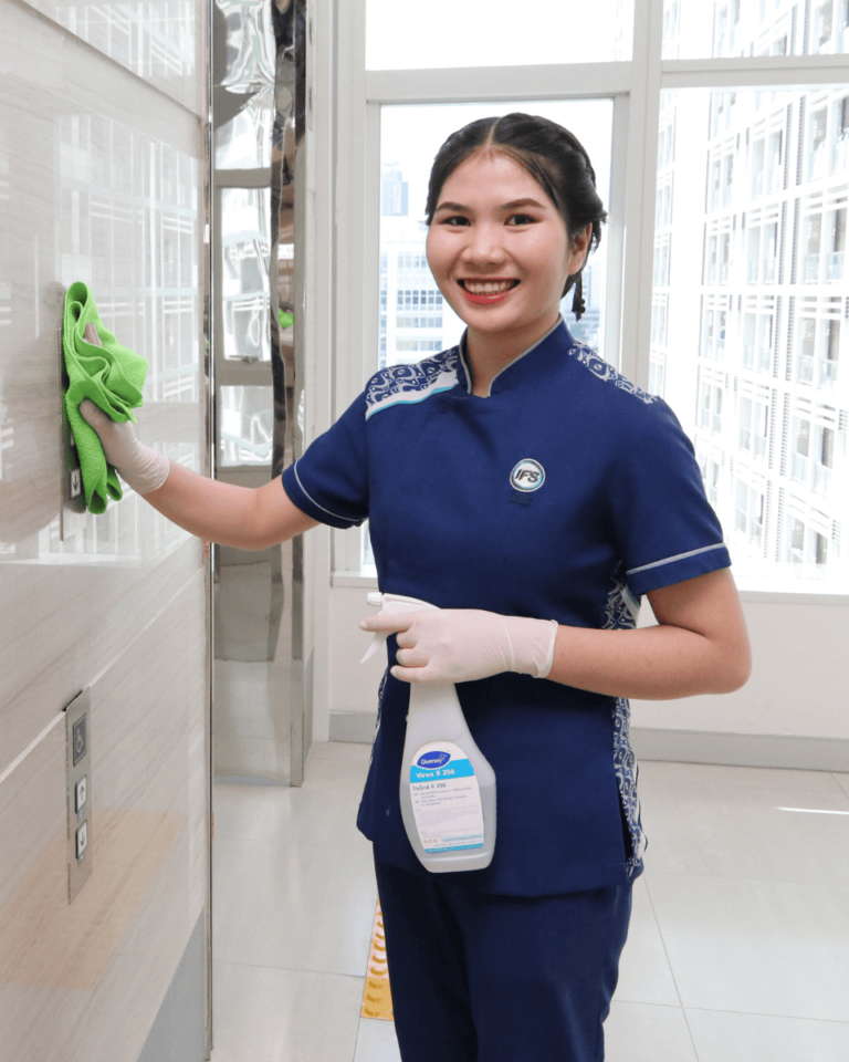 พนักงานทำความสะอาดของ IFS ยิ้มทักทายขณะเช็ดทำความสะอาดผนัง พร้อมใช้น้ำยาฆ่าเชื้อและถุงมือ สะท้อนถึงงานบริการทำความสะอาดประจำวันอย่างมืออาชีพ